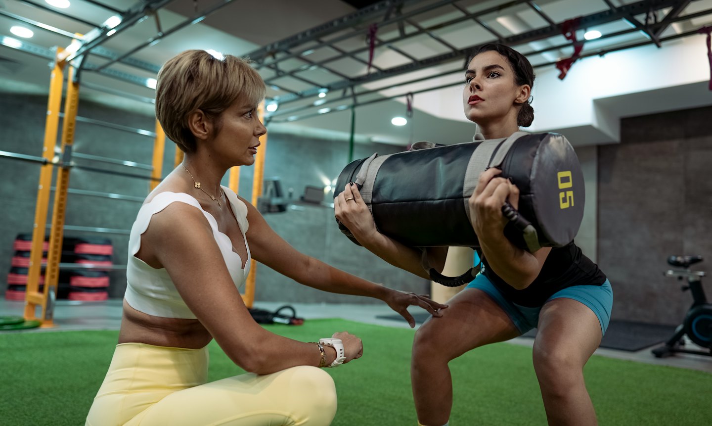 Trainer leitet Frau beim Kniebeugen mit Sandsack im Fitnessstudio an.